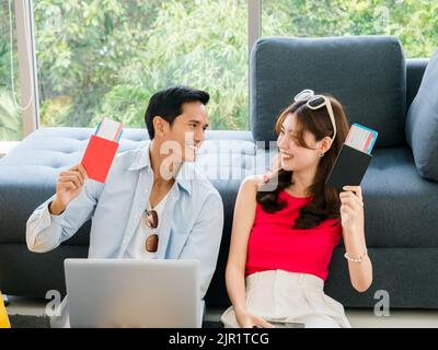 Joyeux couple asiatique prêt à voyager, jeune homme et femme, tenant et montrant passeport tout en utilisant l'ordinateur portable ensemble pour la réservation de billet d'avion, tr Banque D'Images