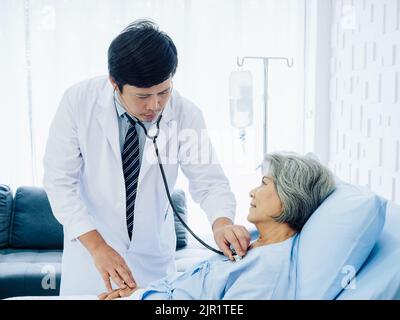 Femme asiatique âgée vêtue de bleu clair, souriant avec joie au lit tandis que le médecin de gentiment masculin en costume blanc utilise le stéthoscope pour l'écouter Banque D'Images