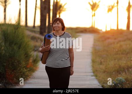 bonne femme d'âge moyen avec tapis de yoga et marcher jusqu'à la plage pour pratiquer le yoga, faire de l'exercice à l'extérieur avec le sentiment de bonheur au lever du soleil le matin. Con Banque D'Images