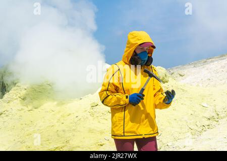 une volcanologue féminine, à l'arrière-plan d'un fumarale fumarate, examine un échantillon de minerai de soufre avec un marteau géologique Banque D'Images