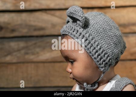 magnifique bébé latina avec la peau brune, distrait regardant d'un côté, avec son visage dans le profil montrant son côté gauche, crochet cap sur un dos en bois Banque D'Images