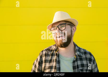 Portrait de rire jeune homme hipster souriant sur le mur jaune magasin arrière-plan extérieur - joli gars à barbe tendance avec chapeau et lunettes debout Banque D'Images