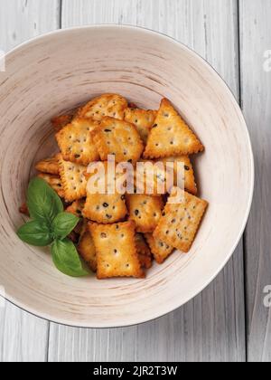 Quelques crackers de saltine dans un bol sur fond de bois blanc. Papillotes salées prêtes à manger avec des graines de sésame noir et blanc. En-cas croquant cuit au four. Banque D'Images