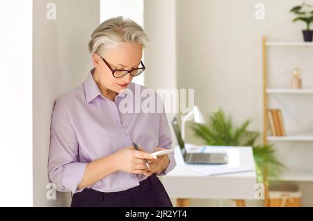 Belle femme d'affaires senior debout au bureau et utilisant son téléphone portable Banque D'Images