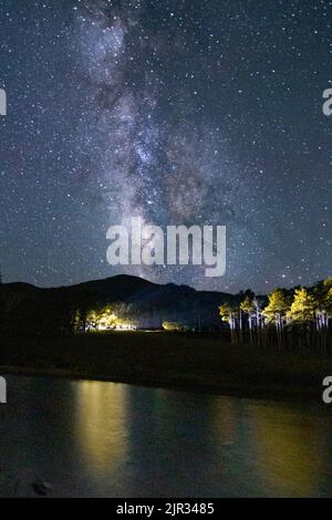 La voie lactée se trouve presque à la verticale au-dessus du lac Isabel dans le sud du Colorado, tandis que les lumières le long de la rive éclairent des peuplements d'épineux Banque D'Images