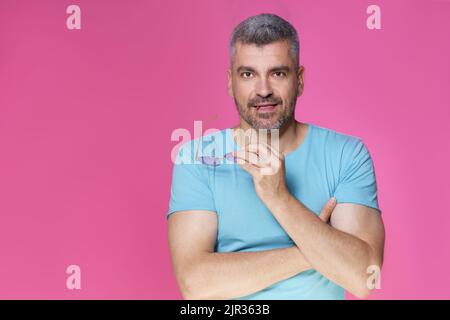 Beau homme adulte avec cheveux gris dans décontracté isolé sur fond rose. Un homme d'âge moyen bien beau debout avec les bras pliés tenant des lunettes de soleil et sourire portant un t-shirt bleu. Banque D'Images