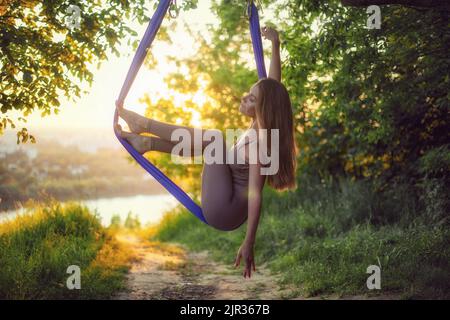 Une jeune gymnaste femelle est engagée dans le yoga aérien, en utilisant une combinaison de poses traditionnelles de yoga, de pilates et de danse en utilisant un hamac au coucher du soleil dans la nature Banque D'Images