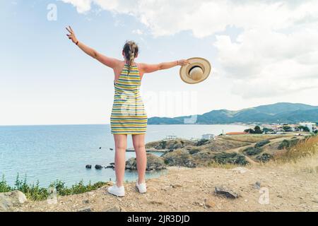 Voyager à l'étranger. Jeune femme blonde caucasienne dans une robe rayée et des sneakers blanches regardant le paysage marin grec à couper le souffle et écartant ses bras dans les deux sens. Photo de haute qualité Banque D'Images