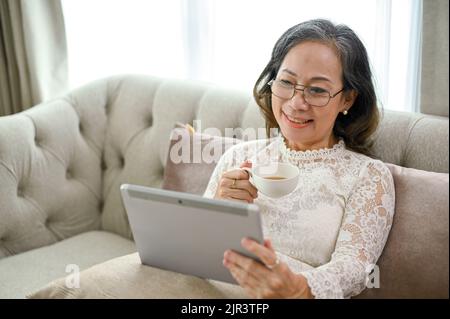 Une femme asiatique d'âge moyen, charmante et attrayante, prend un thé ou un café chaud tout en se relaxant à l'aide d'une tablette numérique dans le salon. Banque D'Images