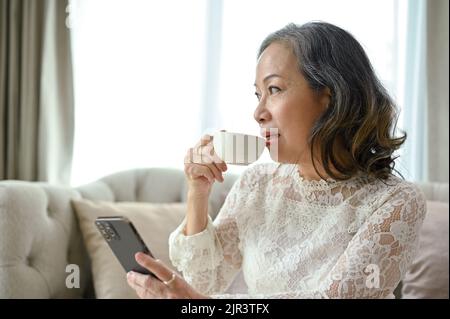 Une femme asiatique d'âge moyen, attrayante et paisible, prend un thé chaud ou un café tout en se relaxant dans le salon. Banque D'Images