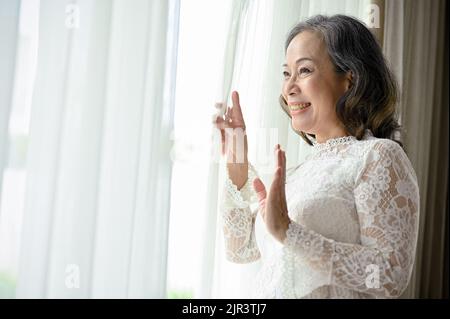 Belle et heureuse femme asiatique d'âge moyen qui regarde par la fenêtre le matin, en admirant la belle vue à l'extérieur de la fenêtre Banque D'Images