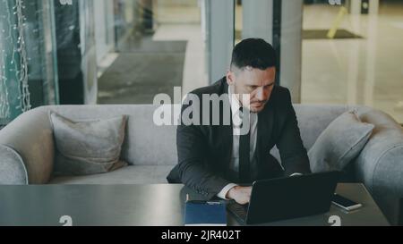 Homme d'affaires caucasien respecté dans un costume noir assis et travaillant concentré sur son ordinateur portable dans un café moderne à l'intérieur Banque D'Images