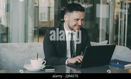 Portrait d'un homme d'affaires caucasien gaie dans des vêtements habillés assis et dactylographiant sur son ordinateur portable tout en écoutant de la musique avec des écouteurs dans ses oreilles dans le café mod. Il sourit et secoue la tête avec joie. Banque D'Images