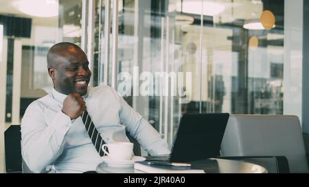 Portrait d'un homme d'affaires afro-américain hilarant dans des vêtements formels souriant, imprimant et travaillant sur son ordinateur portable dans un café aéré pendant la pause déjeuner. Banque D'Images