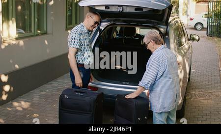 Des aînés joyeux se rendent à la retraite tout en ayant de lourds bagages et un chariot de voyage. Couple senior mettant des bagages dans le coffre de voiture tout en se prépare pour les vacances. Banque D'Images