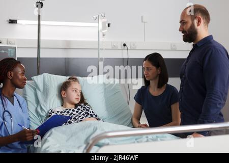 Le personnel de l'établissement de santé pour enfants parle avec les parents de l'état de santé de la fille malade et du programme de traitement. Infirmière assise à côté d'une petite fille malade tout en consultant ses symptômes de maladie. Banque D'Images