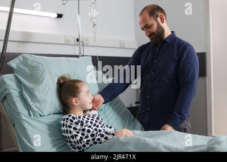 Un père aimant réconfortant une fille malade hospitalisée qui se repose dans le lit du patient dans la chambre de service pédiatrique de l'hôpital. Parent attentionné prenant soin de la petite fille malade tout en touchant son visage. Banque D'Images