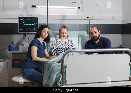 Photo portrait de parents attentionnés assis à côté d'une fille malade dans le lit d'un patient de la clinique de soins de santé d'enfants tout en regardant l'appareil photo. Famille à l'intérieur de la salle pédiatrique de l'hôpital. Banque D'Images