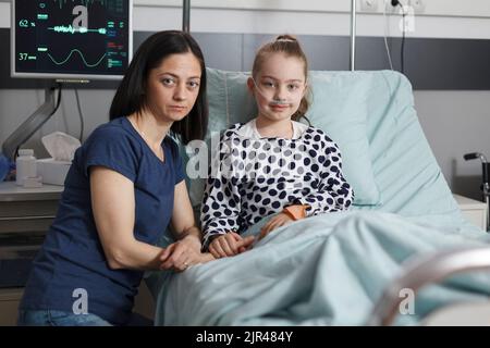 Femme assise à côté d'une petite fille malade dans la chambre d'un établissement de santé pour enfants tout en regardant la caméra. Portrait de la mère soignante réconfortant la fille malade hospitalisée se reposant dans le lit du patient. Banque D'Images