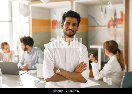 Portrait d'un homme d'affaires indien souriant debout dans un bureau moderne sur l'arrière-plan de ses collègues Banque D'Images