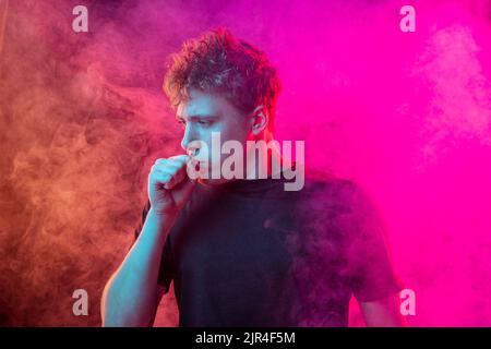 Suffoquer dans la fumée. Jeune homme effrayé avec des émotions stressées isolé sur fond rose avec des nuages de fumée. Concept d'expression faciale Banque D'Images