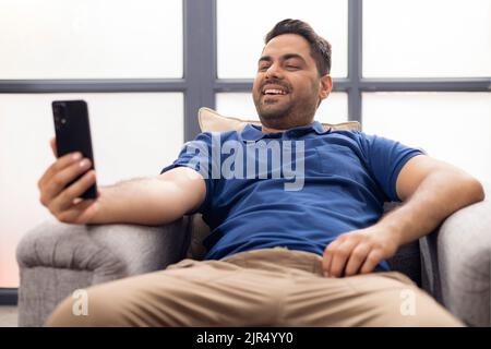 Portrait d'un jeune homme souriant à l'aide d'un téléphone portable assis sur un canapé à la maison Banque D'Images