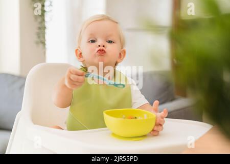 Adorable bébé blond, assis à la table sur une chaise haute avec une cuillère à la main et assiette jaune avec petit déjeuner devant lui, compétences d'entraînement pour se nourrir, porter un bavoir vert. Bébé mangeant par lui-même Banque D'Images