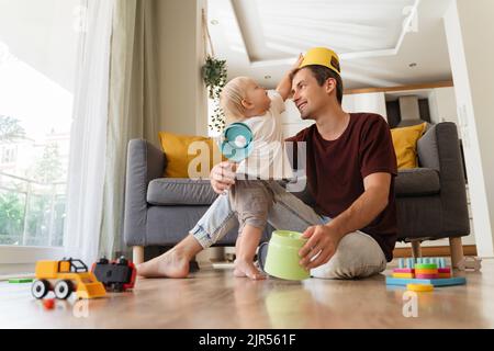 Le jeune père et le fils s'amusent ensemble. Bébé blond tout-petit mettant le jouet sur la tête de papa assis sur le sol dans le salon, papa divertissant son petit garçon, jouant des jouets, des jeux actifs à la maison Banque D'Images