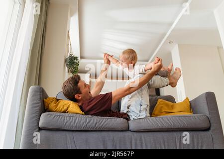 Vue latérale d'un jeune père qui profite du temps avec son fils qui le soulève sur ses jambes, allongé sur un canapé, tandis que sa mère se repose le dimanche. Père jouant avec son tout-petit garçon. Bonne enfance insouciante Banque D'Images