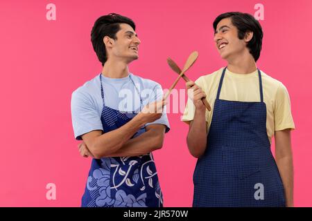 Deux adolescents en tablier de chef debout avec des spatules sur fond rose Banque D'Images