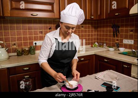 Femme charmante dans une casquette de chef et tablier noir se tient sur une île de cuisine et pèse le sucre sur une balance dans une cuisine maison Banque D'Images