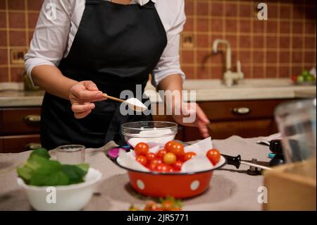 Vue rognée. Femme au foyer dans le tablier du chef noir tenant une cuillère à café en bois avec du sucre tout en préparant des cornichons dans la cuisine maison Banque D'Images