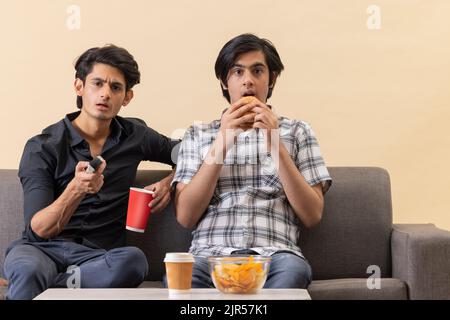 Deux adolescents heureux qui mangent des fast-foods et regardent la télévision ensemble à la maison Banque D'Images