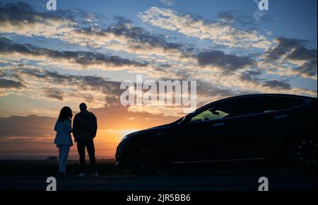 Silhouette de couple romantique qui est arrivé en voiture moderne hors de la ville pour admirer le coucher de soleil sur les champs le soir. Couple de jeunes se tenant main l'un l'autre et regardant la beauté naturelle du coucher de soleil. Banque D'Images