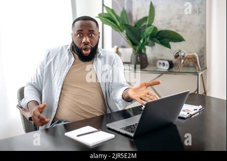 Un homme d'affaires afro-américain stupéfait, un employé de la société assis dans un bureau moderne à une table, sur son lieu de travail, semble surpris par la caméra avec un visage émotif et à yeux larges Banque D'Images