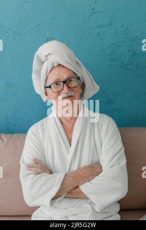 Joyeux homme senior dans des lunettes assis au canapé dans des peignoirs et regardant l'appareil photo. Banque D'Images