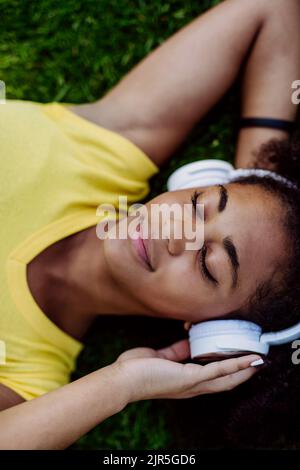 Fille multiraciale couchée dans l'herbe et appréciant la musique dans les écouteurs, vue du dessus. Banque D'Images