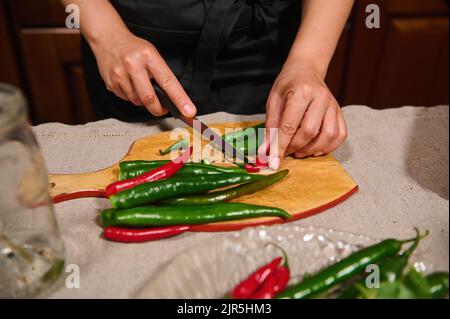 Détails: Mains de la femme de ménage en tablier de chef noir tranchant des piments rouges et verts sur une planche à découper en bois Banque D'Images