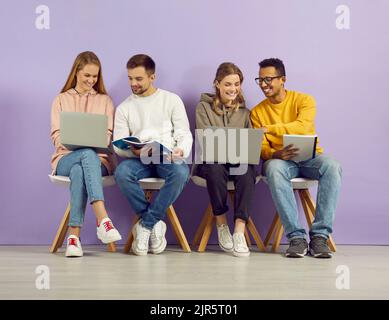 Les étudiants assis en rangée sur fond violet parlent et étudient à l'aide de gadgets et de carnets Banque D'Images