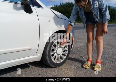 Une femme se penche en posant la main sur un pneu de roue pour vérifier l'état de la voiture blanche garée sur la route Banque D'Images