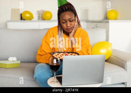 Triste femme afro-américaine dans un chapeau de fête célébrant l'anniversaire en ligne, assis sur un canapé et regardant l'écran Banque D'Images