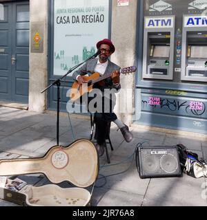 Busker portant un chapeau joue une guitare dans les rues de Porto, au Portugal, avec un haut-parleur et un étui de guitare devant et un distributeur automatique de billets derrière. Banque D'Images