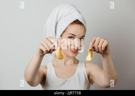 Jeune femme caucasienne tenant dans les mains patchs d'oeil de collagène doré. Isolé sur fond gris de studio. Femme souriante utilisant un cosmétique professionnel pour la procédure anti-âge. Banque D'Images