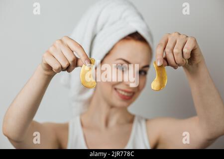 Jeune femme caucasienne tenant dans les mains patchs d'oeil de collagène doré. Isolé sur fond gris de studio. Femme souriante utilisant un cosmétique professionnel pour la procédure anti-âge. Banque D'Images