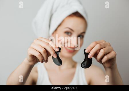 Jeune femme caucasienne tenant dans les mains des timbres noirs d'oeil de collagène. Isolé sur fond gris de studio. Femme souriante utilisant un cosmétique professionnel pour la procédure anti-âge. Banque D'Images