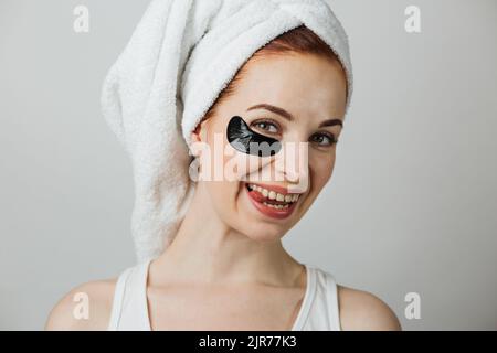 Portrait d'une jeune femme agréable dans une serviette posant en studio avec un timbre noir de collagène sous l'œil montrant sa langue. Charmante femme utilisant des cosmétiques pour les soins de beauté. Banque D'Images