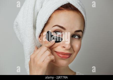 Gros plan sur le visage de la jeune femme adorable, dans une serviette après la douche, en utilisant le patch de collagène noir pour lisser la peau sous les yeux. Concept soins de beauté. Banque D'Images