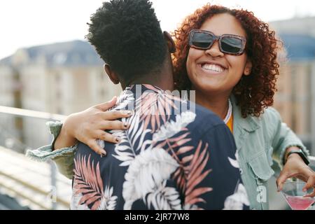 Jeune femme noire gaie dans des lunettes de soleil donnant un câlin à l'homme afro-américain tout en appréciant la fête sur le toit Banque D'Images