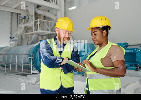 Deux ingénieurs interculturels en réseaux de casques de travail et de casques de sécurité ou en regardant par le biais d'un schéma électronique ou d'un plan de construction Banque D'Images