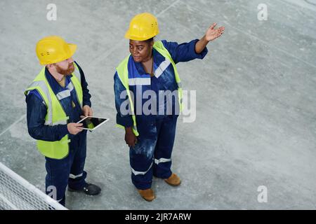 Jeune femme confiante en casque et en uniforme montrant de nouveaux équipements de machines à un collègue pendant la discussion lors de la réunion Banque D'Images